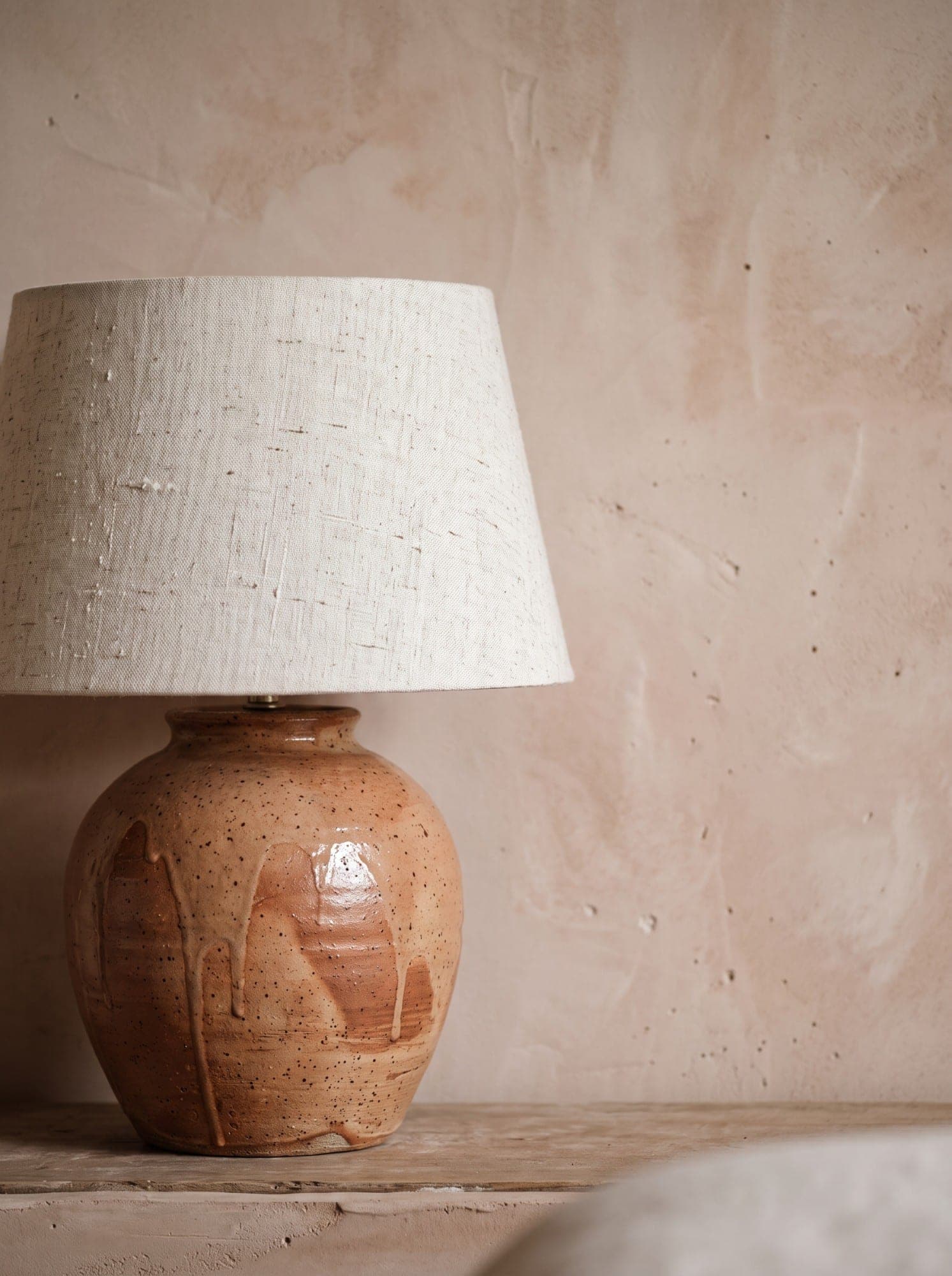 A speckled terracotta pottery lamp with a linen shade against a blush plaster wall